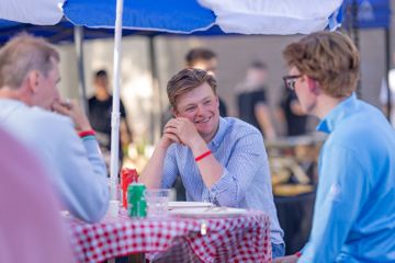 A young St Andrews alumnus sitting with friends at a picnic table, covered with a red and white checked cloth and with a parasol above.