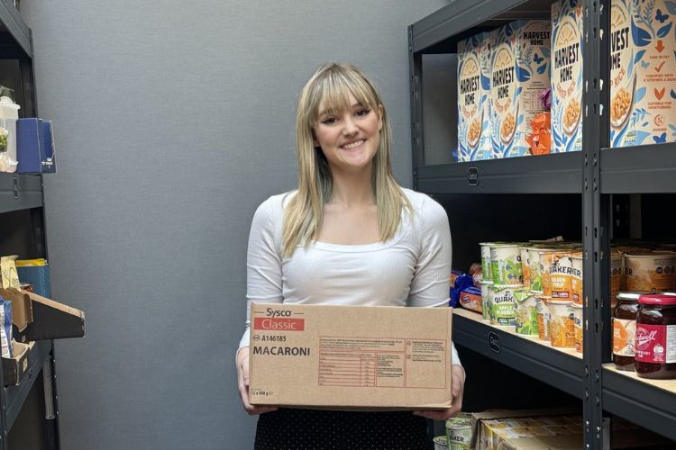 A female student faces the camera holding a packing box labelled macaroni. She is surrounded by shelving units stocked with storecupboard items.