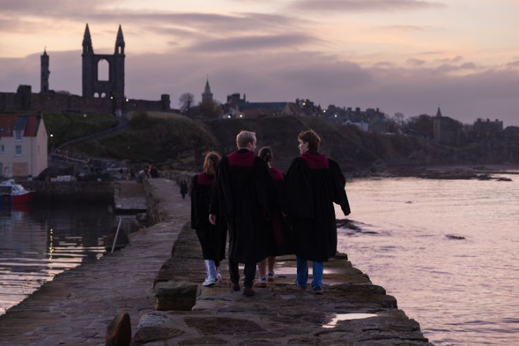 A small group of students in black and burgundy postgraduate gowns walk down the pier away from the camera.