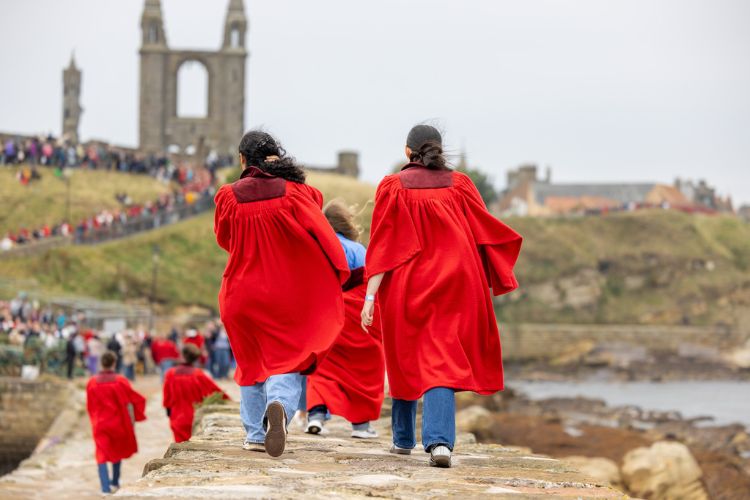 Two female undergraduate students wearing red gowns, pictured walking away down a stone pier towards historic cathedral ruins.