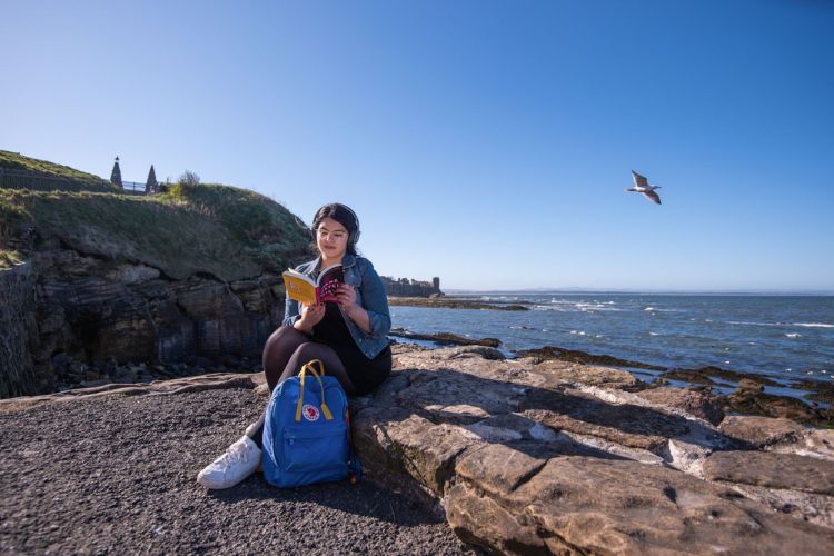 Student reading a book sitting on a rock at St Andrews harbour