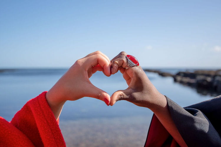 Two students at the University of St Andrews make a heart shape with their hands by the sea.