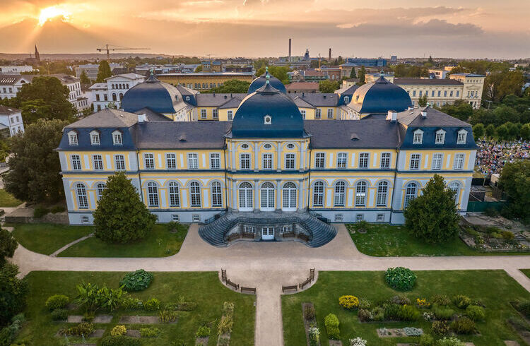 An elegant Baroque-style palace with a symmetrical façade, painted in soft yellow and white tones. Formal botanic garden in the foreground.