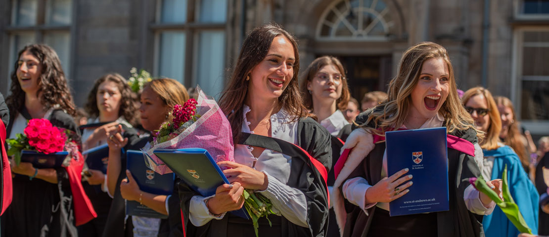 Two happy graduates with more happy graduates in the procession behind them in St Salvator's Quad