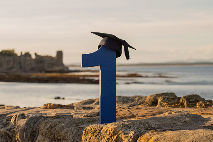 A statue of the number '1' on the pier at St Andrews, with a mortarboard on top and a view of the harbour in the background