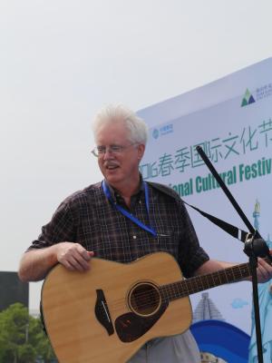 Professor Snow stood in front of a sign advertising a Cultural Festival, playing a guitar