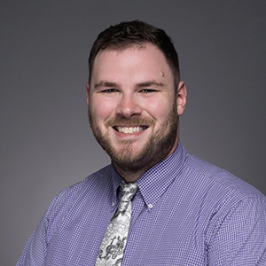 Mitchell, looking to camera in a 3/4 pose wearing a blue checked shirt and grey tie.