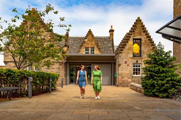 The outside of the Wardlaw Museum on a summer's day. Two females walk towards the camera smiling.