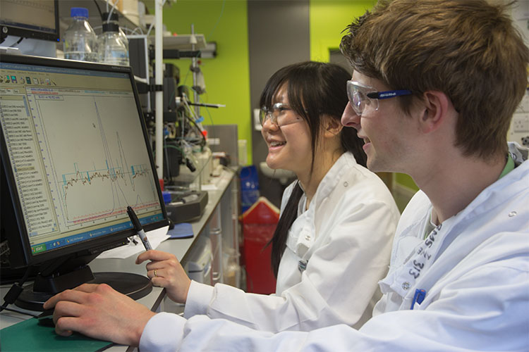 Two students in white lab coats looking at data on a computer
